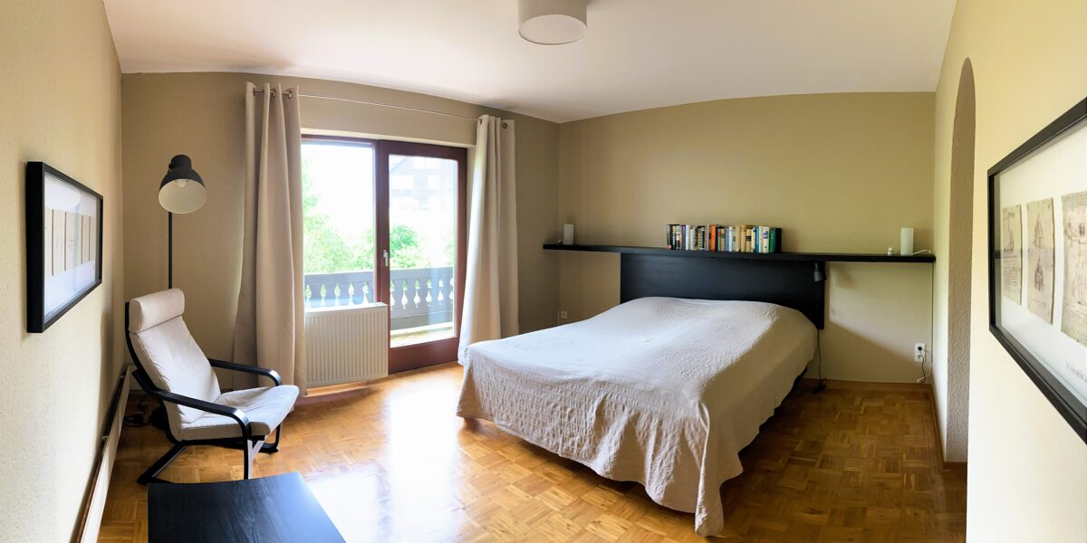 Gruppenunterkunft Tannenhaus - Blick auf das Bett in einem unserer Doppelzimmer mit anschließendem Balkon, Bücherreal, Wandbild, Vorhängen und Leseecke.